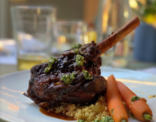 A plated lamb shank with sauce, served on couscous and accompanied by two carrots, garnished with green sauce.