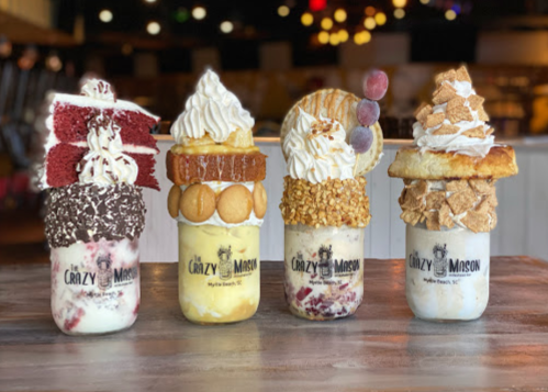 Four extravagant milkshakes topped with whipped cream, cookies, and various sweets, displayed on a wooden table.