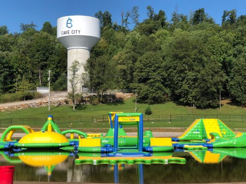 A water park with inflatable attractions on a lake, featuring a water tower labeled "Cave City" in the background.