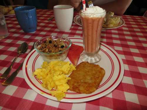 A breakfast plate with scrambled eggs, hash browns, fruit, granola, and a milkshake on a checkered tablecloth.