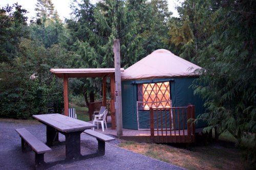 A cozy yurt surrounded by trees, featuring a wooden deck and picnic table in a serene outdoor setting.