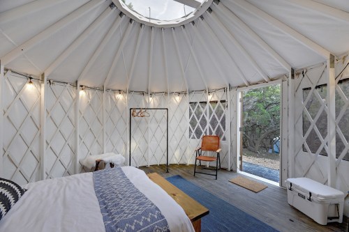 Cozy interior of a yurt with a bed, chair, and natural light from a circular skylight.