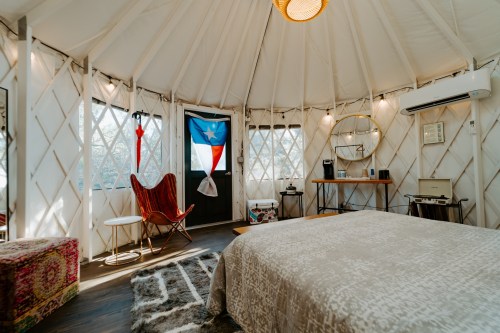 Cozy interior of a yurt with a bed, chair, and decorative elements, featuring large windows and warm lighting.