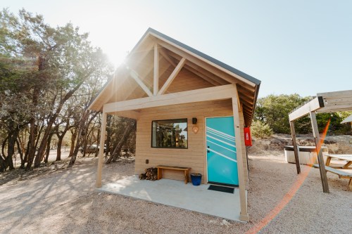A small, modern cabin with a blue door, surrounded by trees and sunlight, featuring a porch and a bench.