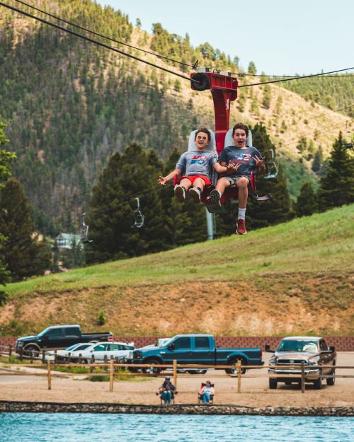 Two children ride a zip line over a scenic landscape with trees and a lake, enjoying a sunny day outdoors.