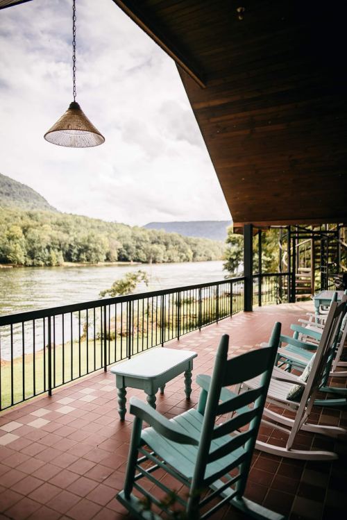 A scenic view from a porch with rocking chairs overlooking a river and lush green hills.