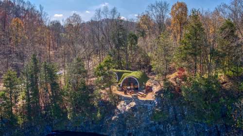 A unique, moss-covered structure nestled among trees on a rocky cliff, surrounded by autumn foliage.