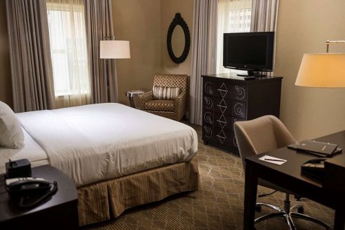 A cozy hotel room featuring a bed, armchair, TV, desk, and decorative mirror with soft lighting.