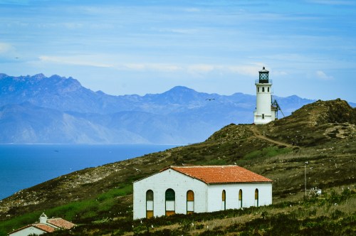 A white lighthouse on a hill overlooking the ocean, with mountains in the background and a small building nearby.