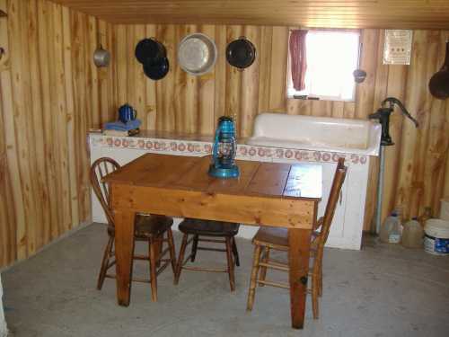 A rustic interior with a wooden table, chairs, and a vintage lantern, surrounded by wooden walls and a sink.