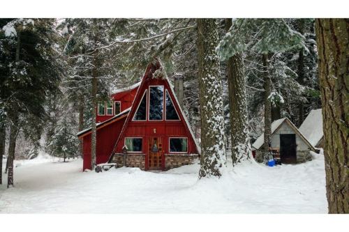 A red A-frame cabin surrounded by snow-covered trees in a winter forest setting.