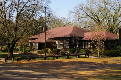A rustic log cabin with a red roof, surrounded by trees and a grassy area, under a clear blue sky.