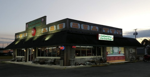 A cozy ice cream shop with a wooden exterior, illuminated at dusk, featuring an "Open" sign and outdoor seating.