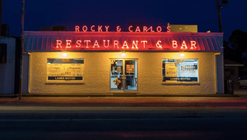 A brightly lit restaurant and bar with a neon sign reading "Rocky & Carlo's" at night.