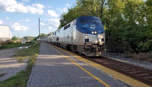 An Amtrak train approaches a station, surrounded by greenery and under a blue sky with scattered clouds.