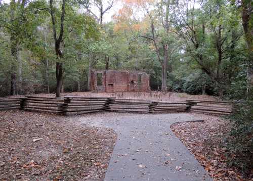 A path leads to the ruins of a brick structure, surrounded by trees and a wooden fence in a forested area.