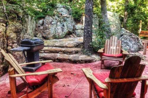 A cozy outdoor seating area with two wooden chairs, a grill, and rocky landscaping surrounded by trees.