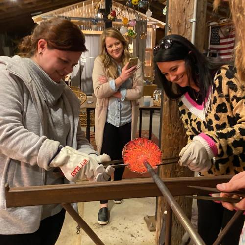 Two women work on glassblowing, while a third woman observes in a cozy workshop setting.