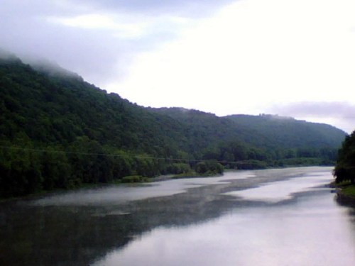 A serene river flows through lush green hills under a cloudy sky, reflecting the landscape in calm waters.