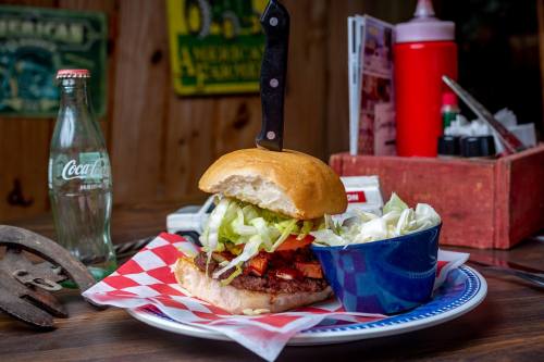 A burger with lettuce, tomato, and a pickle, topped with a knife, served with a side of coleslaw and a vintage soda bottle.