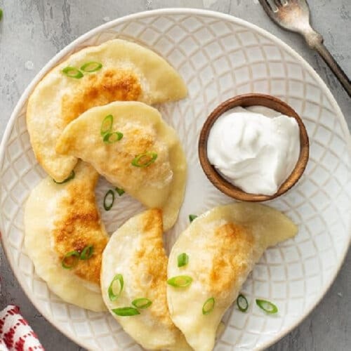 A plate of golden-brown dumplings garnished with green onions, served with a small bowl of sour cream.