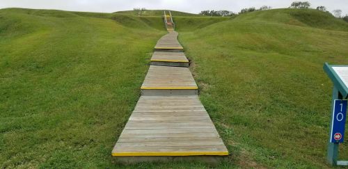 A wooden walkway leads through grassy hills, with a set of stairs visible in the distance.