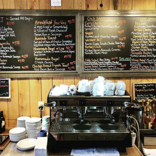 A coffee machine in front of a menu board featuring breakfast, sandwiches, and salad options on a wooden wall.