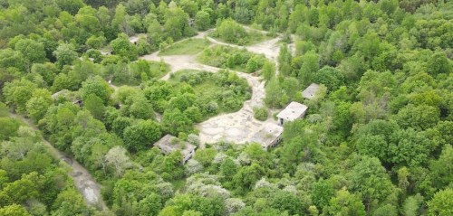 Aerial view of overgrown land with scattered abandoned buildings surrounded by dense green trees.