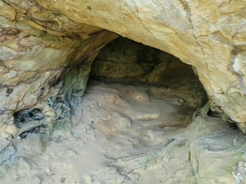 A rocky cave entrance with uneven walls and sandy ground, surrounded by natural stone formations.