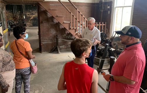 A group of people listens to a guide in a workshop, surrounded by tools and machinery, with a staircase in the background.