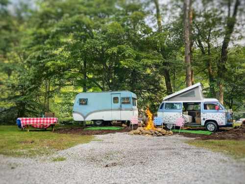 A vintage camper and van parked in a wooded area, with a picnic table and a campfire in front.
