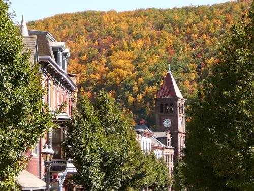 A charming street lined with trees, leading to a clock tower and vibrant autumn foliage on the hills in the background.