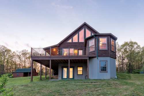 A modern wooden house with a deck, large windows, and a scenic backdrop of trees at dusk.