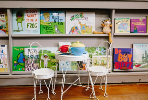 A small table with two chairs, set with a teapot and cups, in front of a colorful bookshelf filled with children's books.