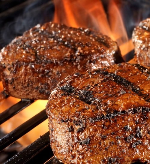 Close-up of juicy steaks grilling on a barbecue, with grill marks and a smoky background.