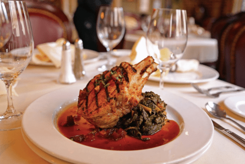 A beautifully plated pork chop on a bed of greens, garnished with herbs, set in an elegant dining setting.