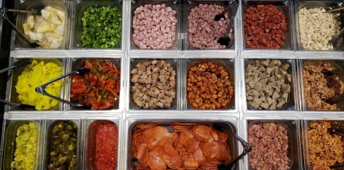 A variety of toppings in containers, including meats, vegetables, and seasonings, arranged in a grid layout.
