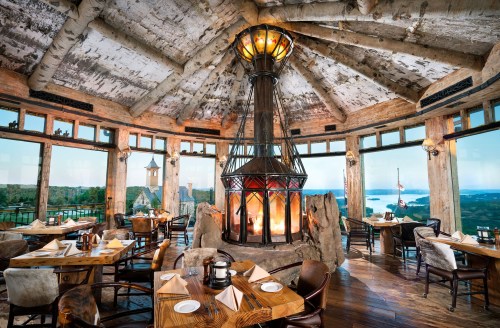 Cozy restaurant interior with a central fireplace, wooden tables, and panoramic windows showcasing a scenic view.