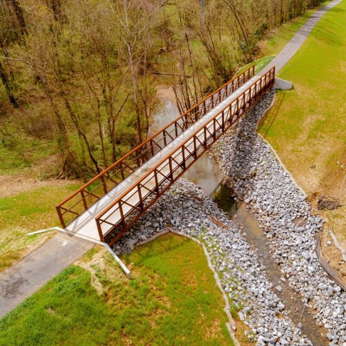 Aerial view of a metal footbridge over a rocky stream, surrounded by green grass and trees.