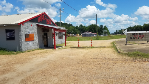 A small barbecue restaurant with a red and gray exterior, surrounded by gravel and open fields under a blue sky.