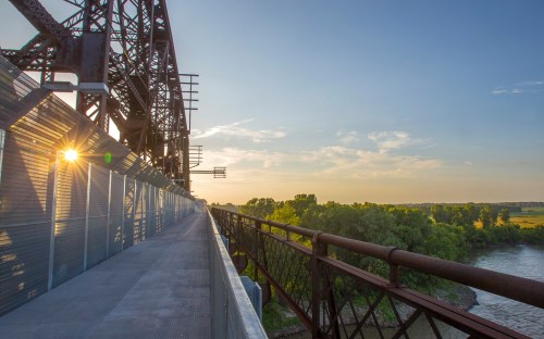 A scenic view of a bridge at sunset, with a river and lush greenery visible in the background.