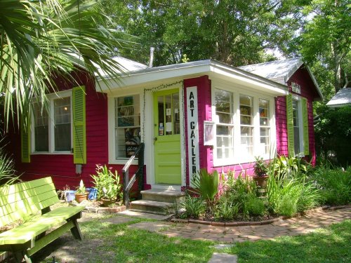 A vibrant pink art gallery with green accents, surrounded by lush greenery and a wooden bench in front.