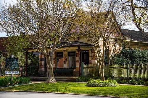 A charming historic house with a porch, surrounded by trees and greenery, and a white picket fence.