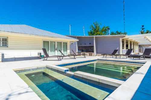 A sunny outdoor area featuring a pool, spa, and lounge chairs beside a modern house. Clear blue sky above.