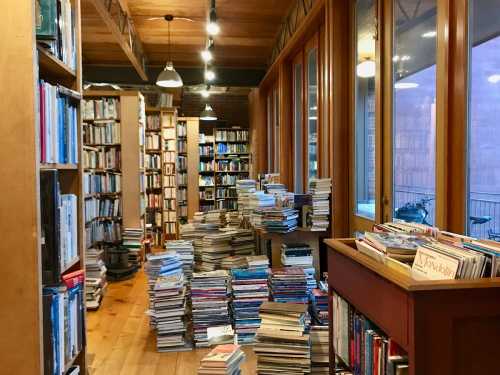 A cozy library with tall bookshelves and stacks of books on the floor, illuminated by warm overhead lights.