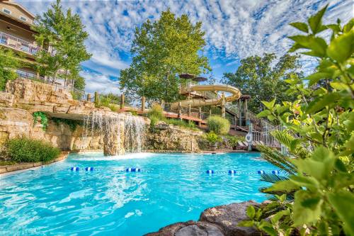 A vibrant pool with a waterfall, surrounded by greenery and a waterslide, under a blue sky with fluffy clouds.