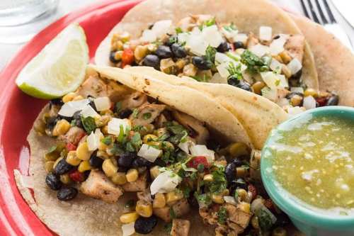 Two tacos filled with chicken, corn, black beans, and onions, served with lime and a side of green salsa.