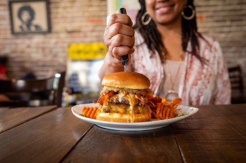 A person holds a fork above a stacked burger with melted cheese and toppings, accompanied by sweet potato fries.