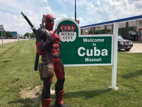 A costumed character resembling a superhero poses next to a "Welcome to Cuba, Missouri" sign.