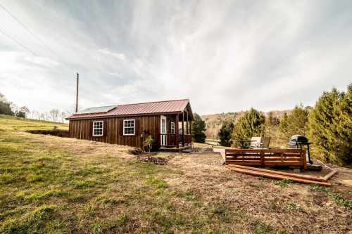 A small wooden cabin with a metal roof, surrounded by grass and trees, featuring a deck and grill area.
